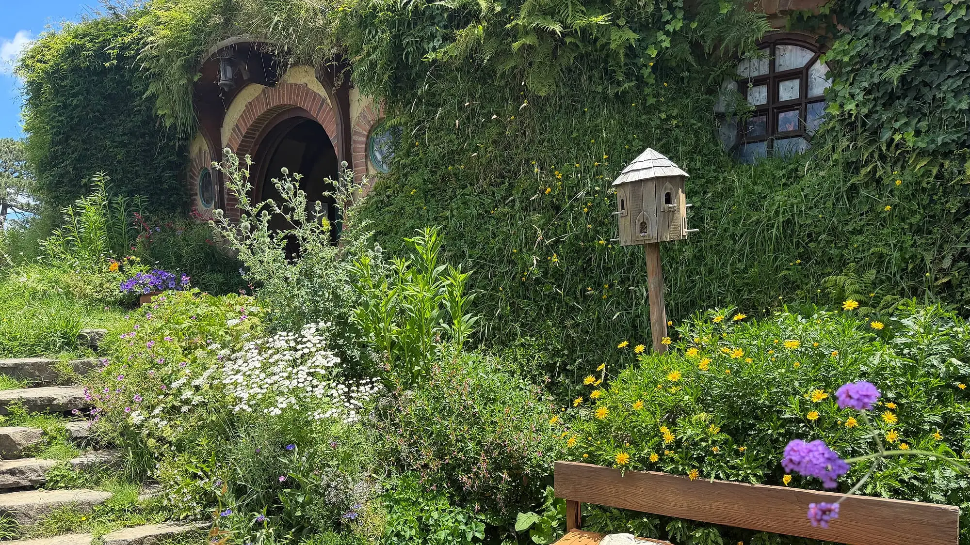 A hobbit-hole entrance at the Hobbiton movie set in the Waikato — round-arched green door framed by ivy and stone steps, wildflower garden with a wooden bench and birdhouse, 90 minutes from Hamilton, New Zealand