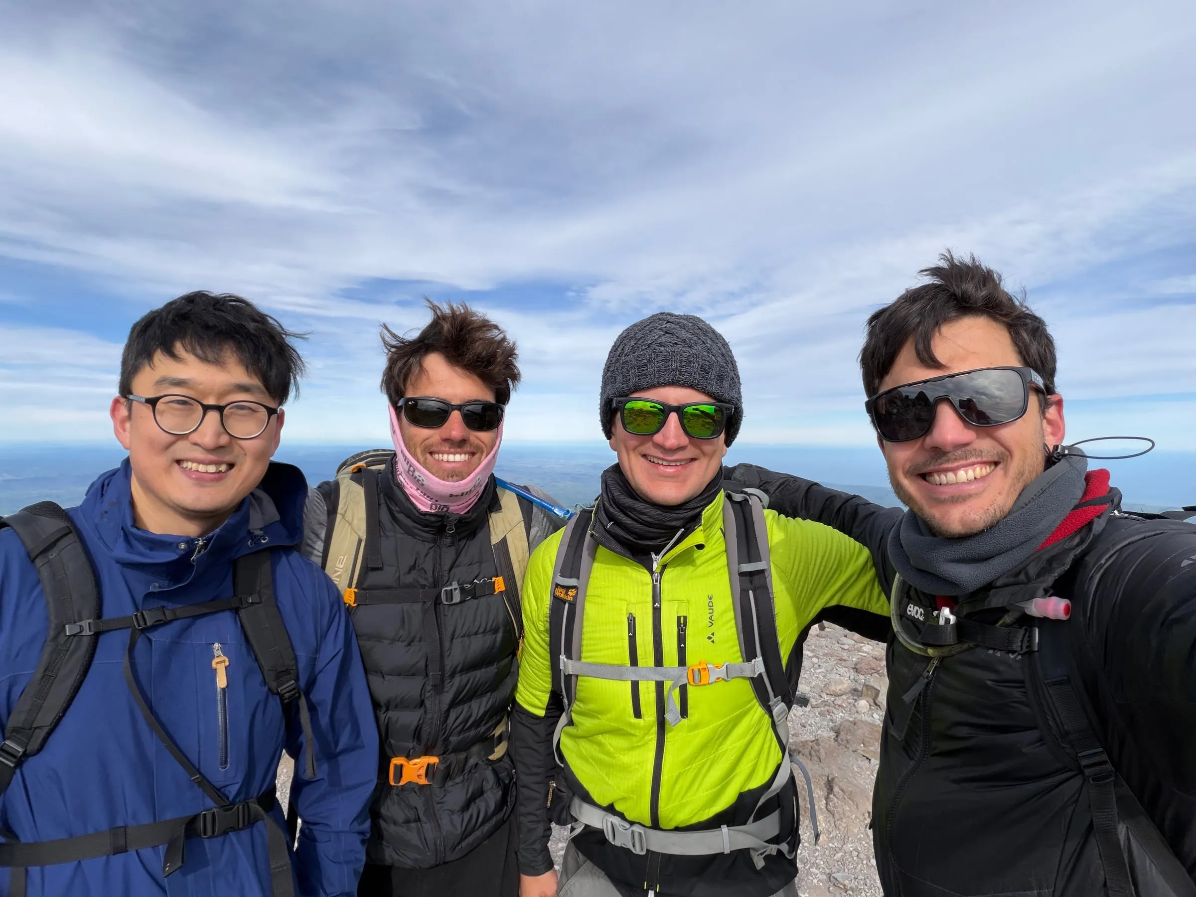 Four people at the summit of Mt Taranaki, smiling for a group photo with the cloud-covered North Island below — from left: Jun Kwon, a friend, Mateusz Gładysz, Juan Gadler