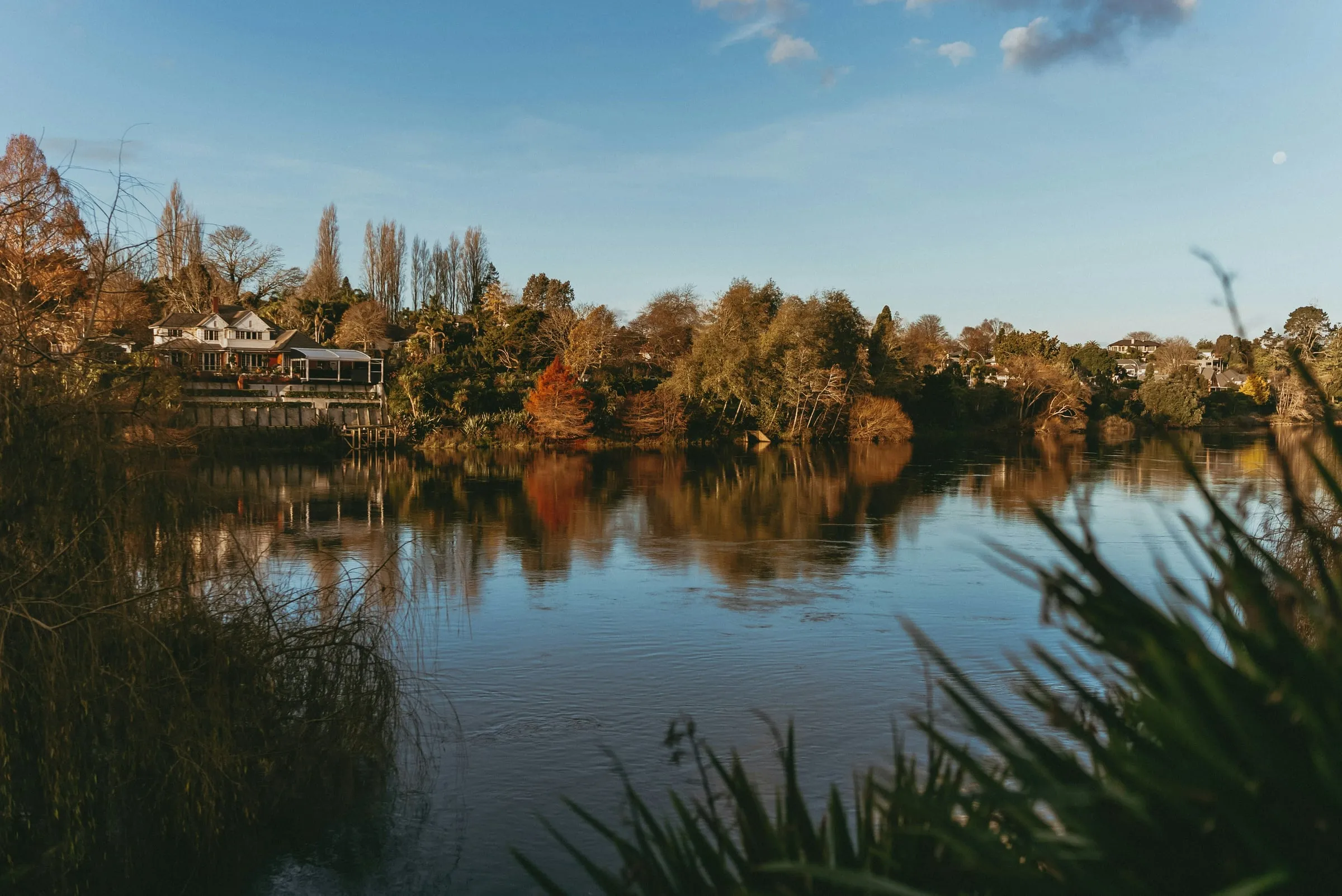 The Waikato River at golden hour, reflecting autumn trees and a riverside Cambridge home