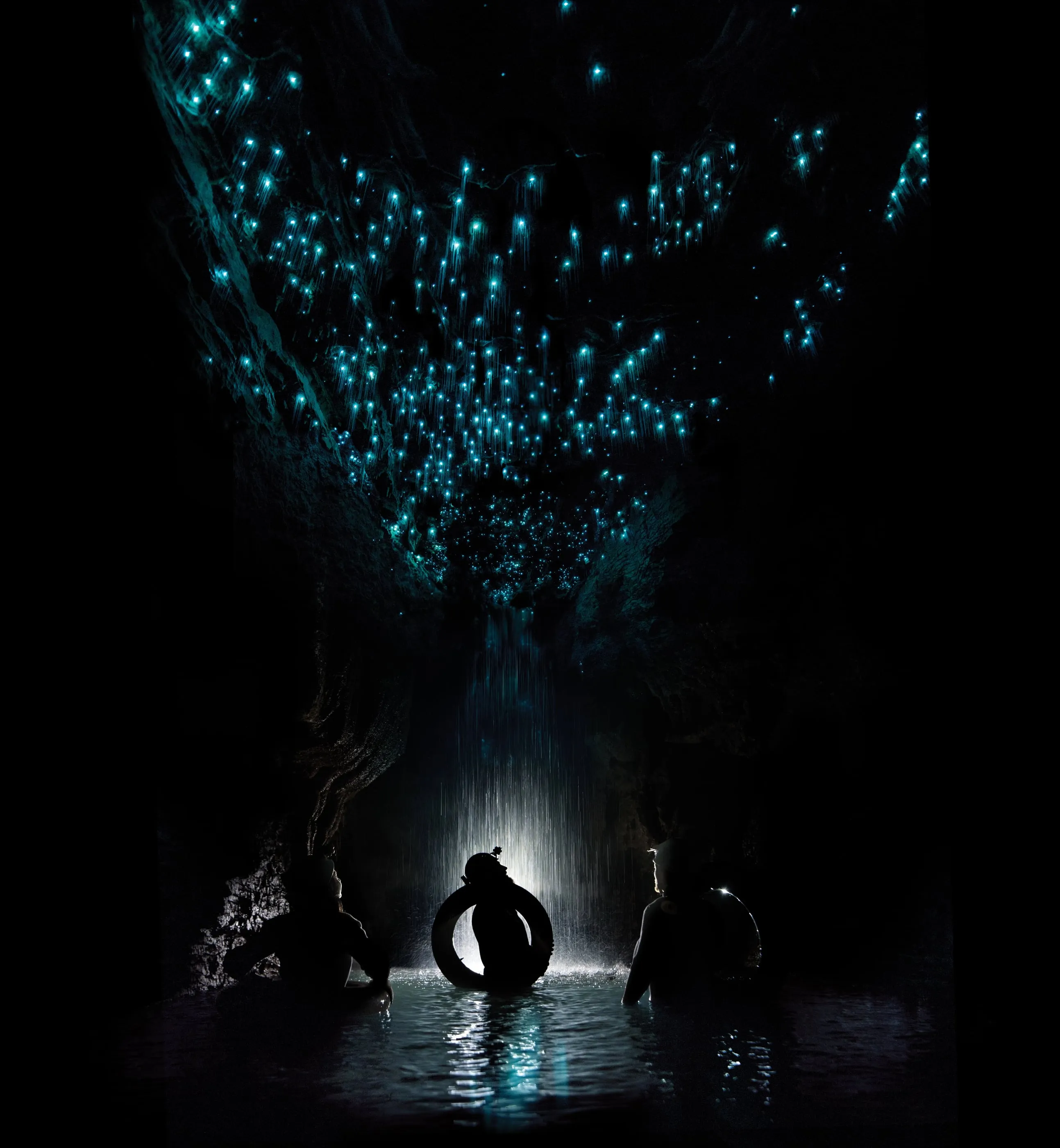 Glow-worms light the ceiling of a Waitomo cave as three figures drift below on inner tubes — Waitomo is an hour from Hamilton, New Zealand